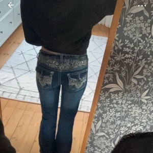 Blå jeans med spetsdetaljer - Snygga blå jeans med unika spetsdetaljer och paljetter på bakfickorna. De har en låg midja och bootcut-stil som ger en trendig look. Perfekta för att lägga till lite extra stil i din garderob.