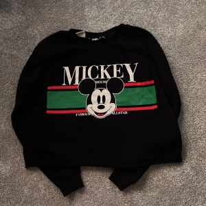 Svart tröja med Mickey Mouse från FB Sister - Säljer en svart tröja med ett stort tryck av Mickey Mouse på framsidan. Tröjan har långa ärmar och en färgglad randig detalj i grönt och rött. Perfekt för Disney-fans! 🖤