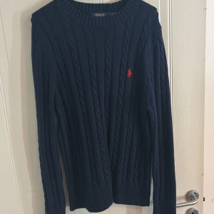 Mörkblå stickad tröja från Polo Ralph Lauren - Säljer en stilren mörkblå stickad tröja från Polo Ralph Lauren med det klassiska röda logotypen på bröstet. Tröjan har en ribbad struktur och långa ärmar, perfekt för kyligare dagar.