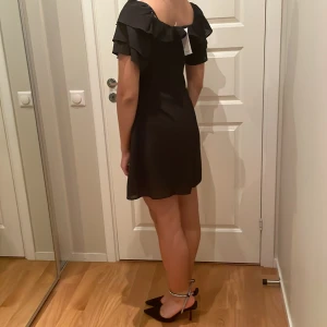 Svart klänning med volangdetaljer - Säljer en elegant svart klänning med volangdetaljer och korta ärmar. Klänningen har en offshoulder-design och en söt rosett i ryggen. Perfekt för en stilren look. Storlek S. Aldrig använt