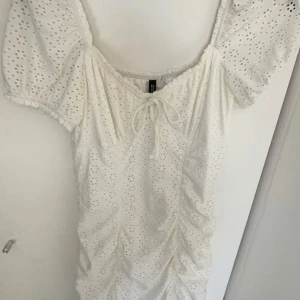 Vit kortklänning - Säljer en söt vit kortklänning med spetsdetaljer och puffiga kortärmar. Klänningen har en v-ringad design med knytning framtill och ett åtsittande passform. Använd några få gånger! 