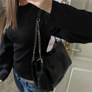 Snygg svart axelväska i skinn med stiliga kedjedetaljer på axelremmen. Perfekt för att ge din outfit en edgy touch. Väskan har en praktisk storlek för att rymma dina nödvändigheter.