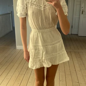 Vit kortklänning med spetsdetaljer - Charmig vit kortklänning med spetsdetaljer och puffiga kortärmar. Klänningen har en elastisk midja och volangkant nedtill. Perfekt för sommaren och studenten!