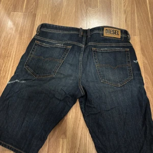 Mörkblå jeansshorts från Diesel - Säljer ett par mörkblå jeansshorts från Diesel med slitna detaljer och klassisk femficksdesign. Shortsen har en slim passform och är perfekta för en avslappnad stil. Märkeslogga på baksidan ger en extra touch. Storlek 32 