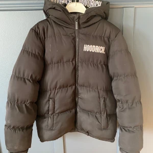 Hoodrich jacka  - Säljer en svart pufferjacka från Hoodrich med huva och dragkedja. Jackan har långa ärmar och två fickor framtill. Perfekt för kyligare dagar. Hoodrich-loggan syns tydligt på bröstet och insidan av jackan.