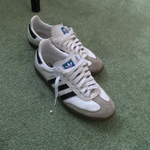 Adidas Samba sneakers - Klassiska Adidas Samba sneakers i vitt och svart med grå detaljer. Skorna har den ikoniska tre-randiga designen och är tillverkade i skinn och mocka. (Storlek 39 1/3)