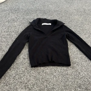 Svart ribbad tröja från Bershka - Säljer en svart ribbad tröja från Bershka med en unik krage och långa ärmar. Perfekt för en stilren look. Tröjan är tillverkad i Bangladesh och har en bekväm passform.