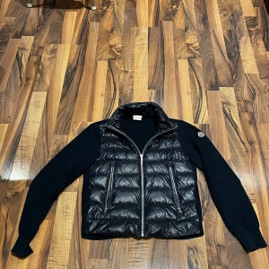 Svart dun jacka  med stickade ärmar från Moncler - Säljer en snygg svart dunjacka från Moncler med stickade ärmar. Jackan har en dragkedja framtill och är perfekt för kyligare dagar och varmare. Den kombinerar stil och funktionalitet på ett grymt sätt. Såklart äkta 