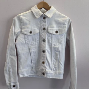 Vit jeansjacka med knappar - Snygg vit jeansjacka med knappar framtill och två bröstfickor med lock. Klassisk krage och långa ärmar ger en tidlös look. Perfekt för vår och sommar!