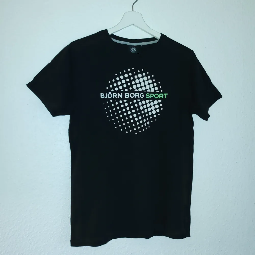 Snygg svart t-shirt från Björn Borg Sport med ett coolt prickmönster och logga i vitt och grönt på framsidan. Perfekt för en sportig look eller avslappnad stil. Gjord i mjukt material för extra komfort.. T-paidat.