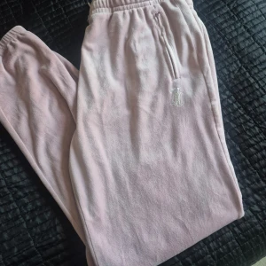 Rosa mjukisbyxor från Juicy Couture - Säljer ett par rosa mjukisbyxor från Juicy Couture med glittrande detaljer. Byxorna har en elastisk midja och dragkedja vid fickorna. Perfekta för en avslappnad stil med en touch av glamour.