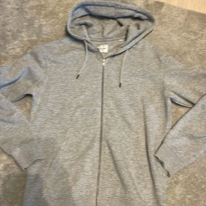 Grå hoodie från Jack & Jones - Säljer en stilren grå hoodie från Jack & Jones. Den har en dragkedja framtill och en praktisk huva med snörning. Perfekt för en avslappnad look.