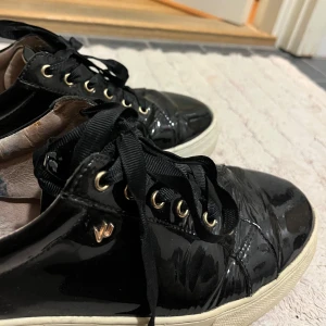 Svarta sneakers med leoparddetaljer - Snygga svarta sneakers i glansigt material med leopardmönstrade detaljer på hälen. Skorna har svarta snören och en vit sula. Storlek 38