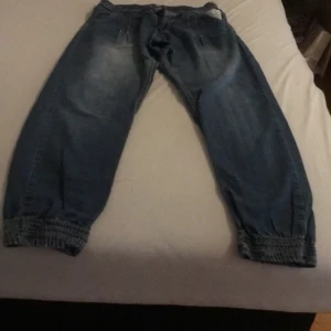 Blå jeans med resårdetaljer - Säljer ett par blå jeans med resår vid bensluten och en avslappnad passform. Byxorna har en klassisk femficksdesign och en dragkedja framtill. Perfekta för en casual look.