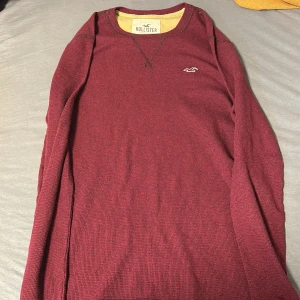 Vinröd långärmad tröja från Hollister - Säljer en vinröd långärmad tröja från Hollister med en liten logga på bröstet. Tröjan har en rund halsringning med en dekorativ söm och är perfekt för kyligare dagar.