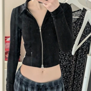 Svart ribbad tröja med dragkedja - Snygg svart ribbad tröja med dragkedja framtill. Tröjan är croppad och har långa ärmar, perfekt för en stilren look. Den har en klassisk krage och är tillverkad i ett bekvämt material.