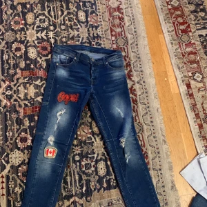 Blå jeans med broderier från Dsquared2 - Snygga blå jeans från Dsquared2 med coola broderier, inklusive en Pepsi-logga och en kanadensisk flagga. Jeansen har en modern design med slitna detaljer och en klassisk femficksmodell. Perfekt för dig som vill sticka ut med stil!