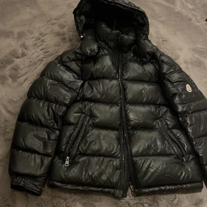 Svart Moncler Maya - Snygg svart pufferjacka från Moncler med huva och dragkedja. Jackan har långa ärmar och praktiska fickor med dragkedja. Perfekt för kyliga dagar. Prissatt billigt pga inget kvitto/låda