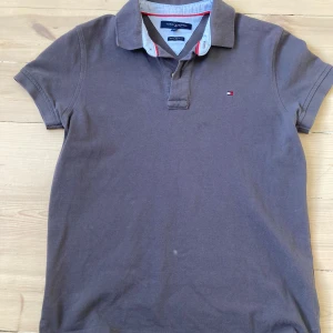 Brun pikétröja från Tommy Hilfiger - Säljer en stilren brun pikétröja från Tommy Hilfiger med klassisk krage och knappslå. Tröjan har korta ärmar och det ikoniska Hilfiger-logotypen broderad på bröstet. Perfekt för en avslappnad men ändå snygg look.