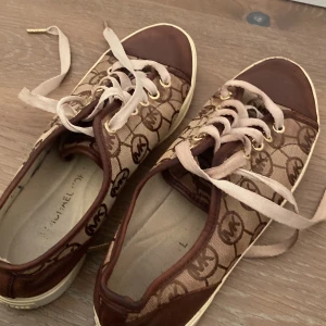 Bruna sneakers från Michael Kors - Snygga bruna sneakers från Michael Kors med deras ikoniska logotypmönster. Skorna har snörning och en bekväm passform. Perfekta för en stilren look.