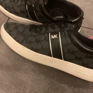 Svarta sneakers från Michael Kors - Snygga svarta sneakers från Michael Kors med ett diskret mönster och märkets logga på sidan. Skorna har en vit sula och snörning framtill. Perfekta för en stilren look.