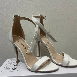 Vita klackskor från Boohoo - Snygga vita klackskor från Boohoo med smala remmar och en elegant design. Perfekta för en stilren look. Klackarna ger en extra touch av elegans och passar utmärkt till festliga tillfällen.