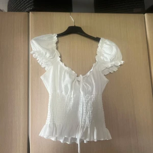 Vit blus med volangdetaljer - Charmig vit blus med volangdetaljer och knytning framtill. Den har en v-ringad design och korta ärmar med volanger. Perfekt för en somrig look.