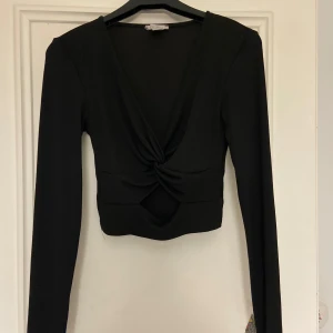 Svart långärmad topp från Misu - Snygg svart långärmad topp från Misu med en djup v-ringning och knytdetalj framtill. Perfekt för en stilren look. Toppen är i ett stretchigt material som ger en bekväm passform.