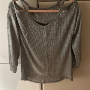 Grå ribbad tröja med cut-out detaljer - Snygg grå ribbad tröja med unika cut-out detaljer vid axlarna. Tröjan har en v-ringad design och långa ärmar, perfekt för en stilren look. Oanvänd 