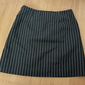 Kjol från Kappahl - Snygg svart kjol med vita ränder från Kappahl. Kjolen har en klassisk design med en normal passform och är perfekt för en stilren look. Den är gjord i ett bekvämt material och har en dragkedja i sidan. Säljer då jag tyvärr inte använder den, aldrig använt och lappar är fortfarande på. Köpt för 350kr