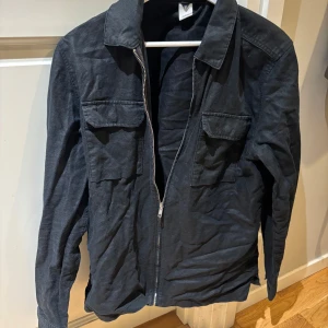 Svart overshirt - Säljer en riktig snygg overshort från Visual Clothing Project. Den i storlek M och är hyfsat tunn, väldigt snyggt med en tröja eller en lätt hoodie under, perfekt till våren eller sommarkvällar. Skriv privat för fler bilder eller funderingar!