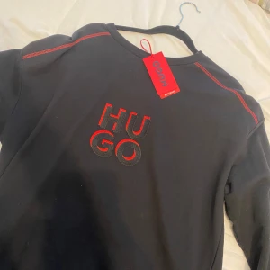 Svart tröja från Hugo - Snygg svart tröja från Hugo med röd logotyp på bröstet och kontrastsömmar. Perfekt för en stilren look. Tröjan har rund hals och långa ärmar.