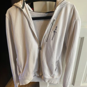 Vit zip hoodie från Sail Racing - Säljer en vit hoodie från Sail Racing med dragkedja och broderad logga på bröstet. Den har en huva och långa ärmar, perfekt för en avslappnad stil. Materialet känns mjukt och bekvämt. Men ett litet hål på framsidan, men det är ihop sytt.