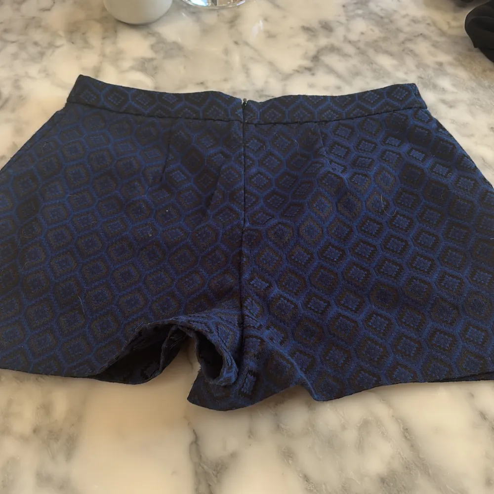 Snygga blå shorts med ett geometriskt mönster i mörkare nyans. De har en dragkedja bak och en stilren design som passar perfekt för en trendig look.. Shortsit.