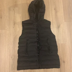 Svart dunväst med huva - Snygg svart dunväst med huva och dragkedja framtill. Perfekt för kyligare dagar och ger en stilren look. Västen har två praktiska fickor med dragkedja.