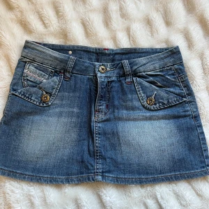 Blå jeanskjol med fickor - Snygg blå jeanskjol med dekorativa fickor fram och bak. Kjolen har en klassisk femficksdesign och knappar med detaljer. Perfekt för en avslappnad stil. Väldigt liten i st för att vara en L skulle säga att den är en S. Midjemått 40cm och längd 33cm