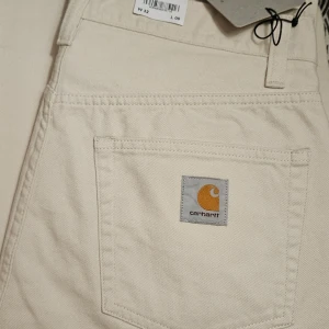 Beige byxor från Carhartt landon pant  - Säljer ett par snygga beige byxor från Carhartt med en loose tapered fit och regular waist. Byxorna har en praktisk dragkedja och är perfekta för en avslappnad stil. Carhartt-loggan syns tydligt på bakfickan.