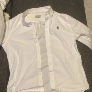 Vit skjorta från Giordano - Säljer en stilren vit skjorta från Giordano. Skjortan har en broderad detalj av ett lejon på bröstet och knappar framtill. Den är slim fit och passar perfekt för en elegant look.