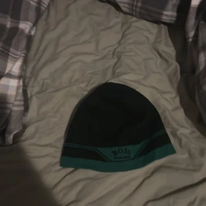 Svart och grön mössa från Hugo Boss - Snygg svart mössa med gröna detaljer från Hugo Boss. Perfekt för att hålla värmen med stil. Märkesloggan är broderad framtill och mössan har en klassisk passform.