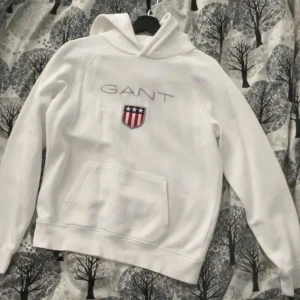 Vit hoodie från GANT - Säljer en stilren vit hoodie från GANT med broderad logga och ett emblem på bröstet. Tröjan har en klassisk känguruficka och är perfekt för en avslappnad stil. Passar för ungdomar runt 15 år.