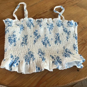 Vit och blå blommig linnetopp från Hollister - Charmig vit linnetopp med blått blommönster. Toppen har smala axelband och en volangdetalj vid nederkanten. 