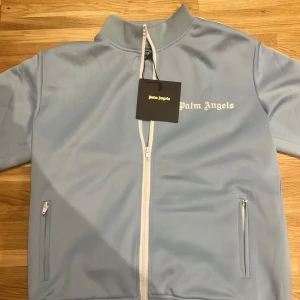 Ljusblå track från Palm Angels - Snygg ljusblå track jacket från Palm Angels med vit dragkedja och vita ränder längs ärmarna. Jackan har en hög krage och långa ärmar, perfekt för en sportig look.