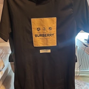 Svart t-shirt från Burberry - Snygg svart t-shirt från Burberry med en stor gul logga på framsidan. Perfekt för dig som vill ha en stilren och lyxig look. T-shirten har en klassisk passform och är kortärmad.