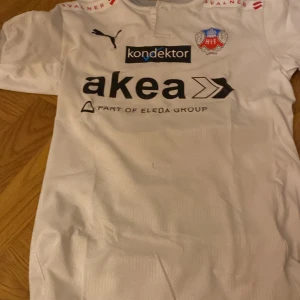 Vit fotbollströja från Puma - Säljer en vit fotbollströja från Puma med korta ärmar. Tröjan har flera sponsorloggor och ett klubbmärke på bröstet. Nummer 11 och namnet 'Svensson' är tryckt på ryggen. Perfekt för fotbollsfans!