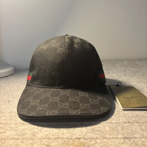 Svart gucci keps - Snygg svart keps från Gucci med diskret mönster och klassiska gröna och röda ränder på sidan. Perfekt för att ge din outfit en lyxig touch. Kepsen har en böjd skärm och justerbar passform. Storlek ”M”