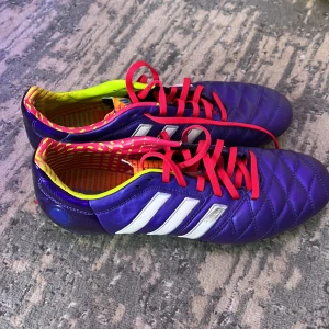 Fotbollsskor från Adidas - Säljer ett par lila fotbollsskor från Adidas med vita ränder och röda snören. Skorna har en quiltad design och en färgglad insida med gula och rosa detaljer. Perfekta för fotbollsplanen! Dom är bara använda nägra få gånger, hör av er vid intresse☺️
