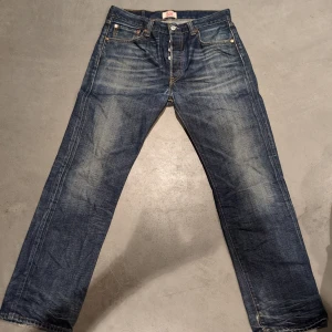 Levis 501 jeans - Klassiska Levis 501 jeans i blå denim med en rak passform. De har en knappgylf och fem fickor. Jeansen har en sliten look med fransade kanter vid bensluten för en cool vintagekänsla.