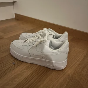 Vita Nike Air Force sneakers - Säljer ett par klassiska vita Nike Air Force sneakers. De har en stilren design med snörning och perforerade detaljer på tån för extra ventilation. Perfekta för en casual look.