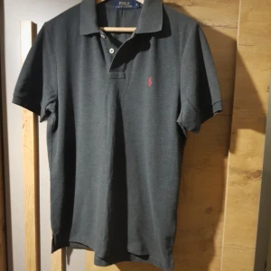 Mörkgrå pikétröja från Ralph Lauren - Snygg mörkgrå pikétröja från Ralph Lauren med klassisk krage och knappslå. Den har en liten röd logga broderad på bröstet. Perfekt för en stilren look.