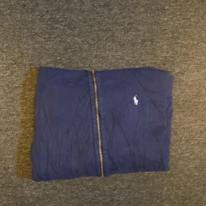 Blå hoodie från Polo Ralph Lauren - Säljer en snygg blå hoodie från Polo Ralph Lauren med dragkedja och klassisk logga på bröstet. Perfekt för en avslappnad stil. Den har långa ärmar och en praktisk huva med snören.